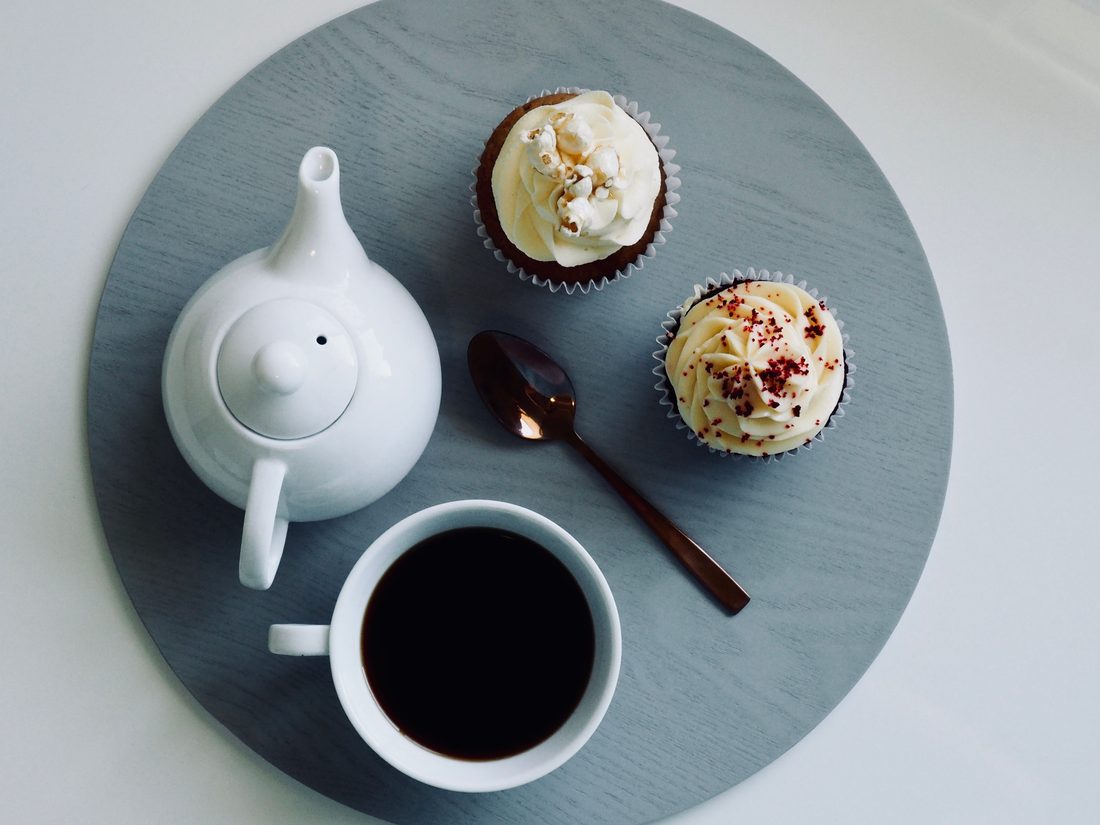 Overhead flatlay of a white teapot, a cup of black coffee, and two frosted cupcakes on a round tray.